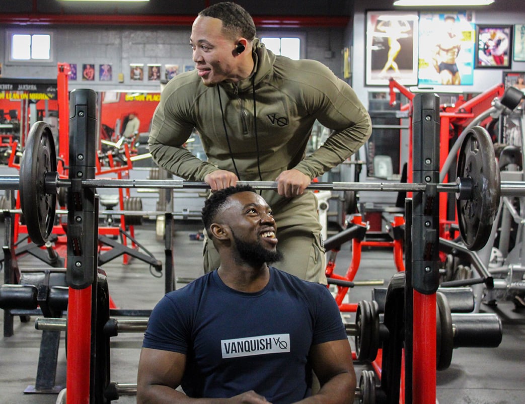 Coach is putting his hand on barbell and stading behind another person who's sitting
